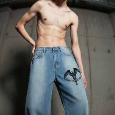 Man wearing light blue jeans with a psalms angels logo, standing against a concrete wall.