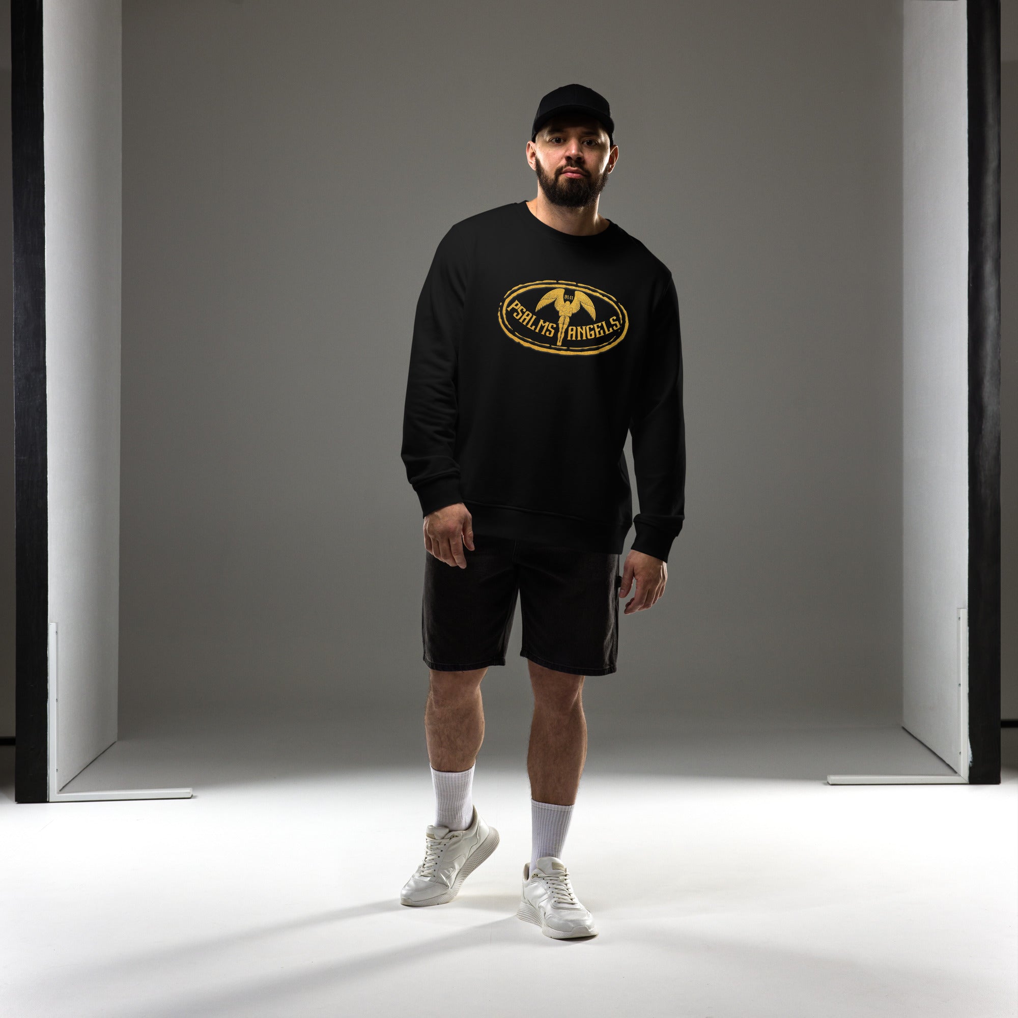 Man wearing a black sweatshirt with a gold Psalms Angels logo and black shorts in a studio setting